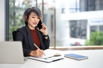 Obraz premium Mature happy joyful confident asian business lady, broker or manager, sitting at table in a modern office, working with laptop, talking to client or employees on smartphone, smiling