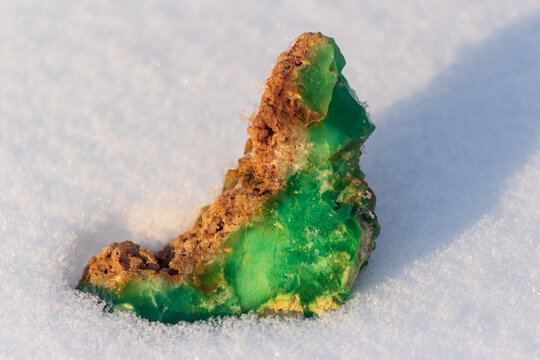 Lush green Kazakhstani chrysoprase sample, an uncut jewel, vibrantly rests on pure white winter snow, showcasing earth's raw, stunning elegance