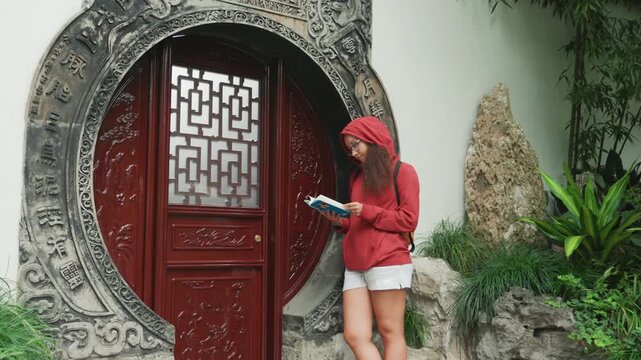 Asian woman phone by moon gate, red hoodie and white shorts beside ornate circular stone doorway, lush plants and rock features, checking smartphone map while pausing on stone steps, calm