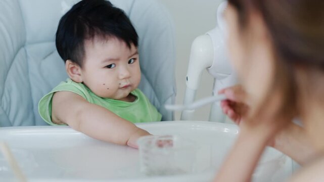 Happy Asian baby boy in high chair smiling at mother while being spoon-fed. Close-up view with natural light in a home setting.