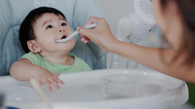 Happy Asian baby boy in high chair smiling at mother while being spoon-fed. Close-up view with natural light in a home setting.