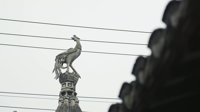 Ornate rooftop phoenix statue against sky, weathered metal crest perched on temple spire, diagonal roof silhouette framing right, layered power wires cutting across scene, overcast mood with muted