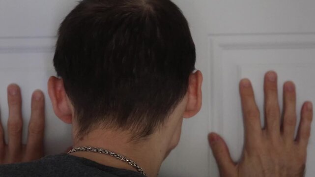 Man carefully approaches a white door and looks through the peephole to check outside. Concept of home security, safety awareness, suspicion, and protection from intruder.