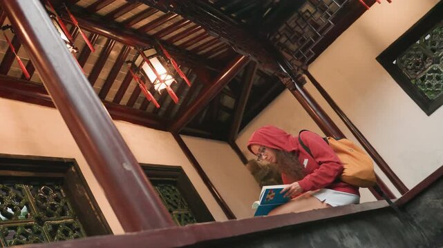 Asian woman in red hijab reading guidebook on wooden veranda near carved windows, warm lantern glow, backpack beside knees, studying traditional patterns, reflective mood, travel student and blogger