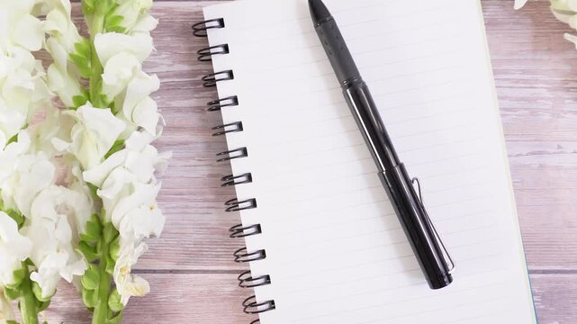 White flowers rest beside a notebook and pen. Wooden table holds these quiet, thoughtful items. Minimalist style feels calm and focused. Perfect for journaling, planning, or creative tasks