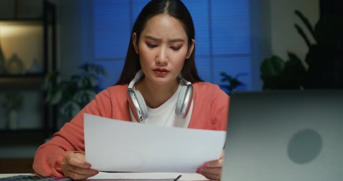 Amid stress, an Asian woman balances her budget and expense sheets, seriously analyzing financial datum on a late-night computer.