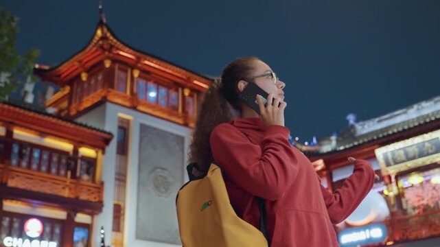 Nighttime lanterned temple exterior, white woman gazing upward. Red hoodie backpack phone call under warm lanterns, ornate pagoda facades and glowing windows, nocturnal urban travel mood, curiosity