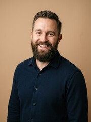 Fototapeta premium A man with a beard and short hair smiles warmly at the camera. He is wearing a dark blue button-down shirt.