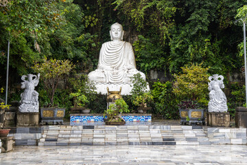Obraz premium A serene white Buddha statue sits in a meditative pose, surrounded by lush greenery at the Marble Mountains in Da Nang, Vietnam. The site is a place of peace and reflection.