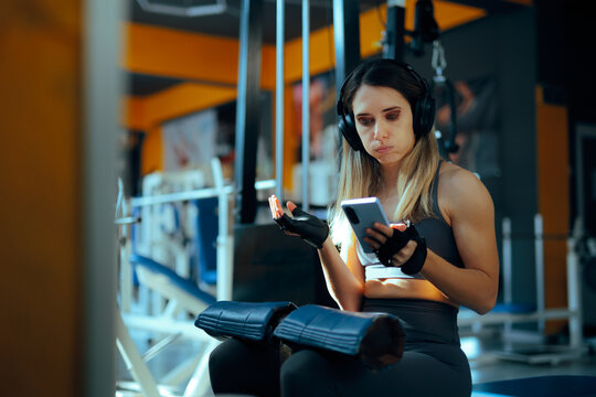 Woman Wearing Headphones checking her Phone in Between Sets. Gym girl being distracted by her phone taking too long breaks to check notification

