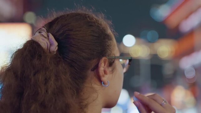 Black student with glasses adjusts earbud in neon city, night street bokeh and soft lights, red hoodie and scrunchie, contemplative profile closeup, smartphone audio connection and quiet commute mood