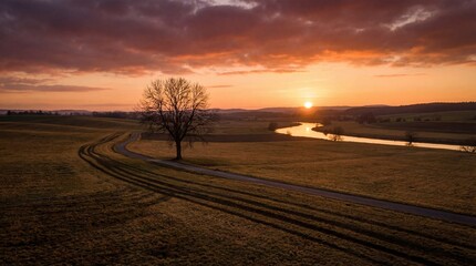 Fototapeta premium A breathtaking sunset paints the sky with fiery hues over a winding river, a solitary bare tree, and a curving dirt path through a vast, tranquil field.