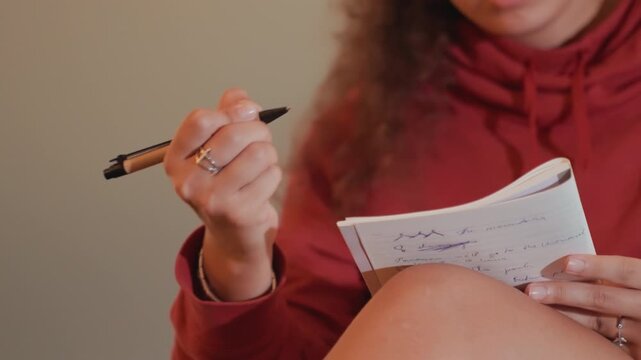 Woman drafting lyrics in notebook with warm bokeh, focused expression in red hoodie, rings and pen visible, pages full of scribbled verses and melody ideas, intimate creative atmosphere