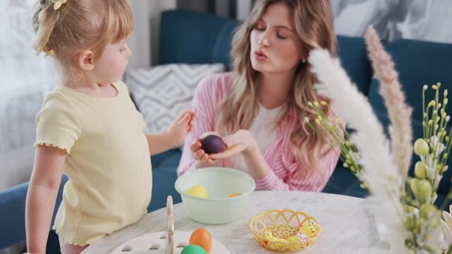 Mother and child decorate Easter eggs together at home. Little girl reaches for egg, smiling with joy. Woman watches warmly, engaged in festive activity