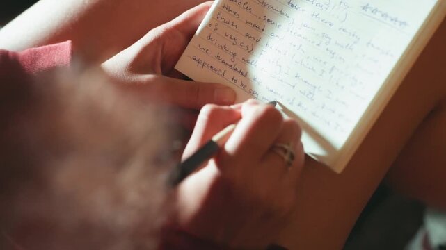 Closeup hands writing in notebook, warm natural light, soft film grain, pen tracing cursive lines, knee resting towel, silver ring on finger, personal diary entries, reflective mood, slow living