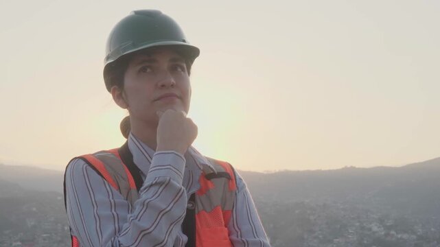 Thoughtful female engineer wearing a safety vest and hard hat looking at the horizon during a beautiful sunrise, representing vision, planning, and professional determination