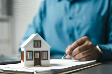 Person signing real estate contract with house model, representing home purchase, property investment, mortgage agreement and financial approval.