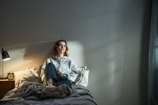 Caucasian young adult woman sitting on bed listening to music with earphones, gazing thoughtfully into distance, expressing calm emotion, morning light casting shadows on wall