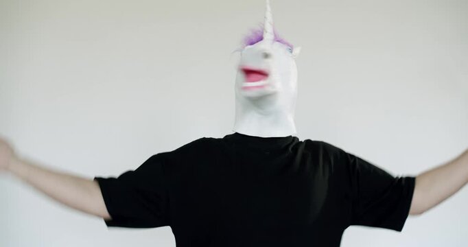 A man wearing a unicorn mask dances and moves around in a studio setting. He is having fun and enjoying himself. The background is plain white, which makes the subject stand out.