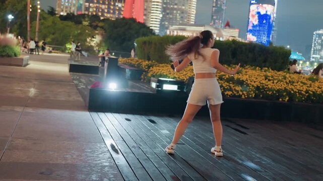Asian woman dancing freely at night city plaza stage jumping spins playful energy sporty sneakers casual outfit illuminated pavement freestyle street performance confident expression