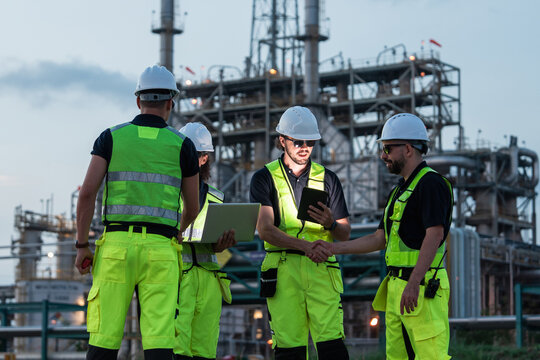 Group of Caucasian industrial engineers wearing safety helmets and reflective uniforms shaking hands at petrochemical plant during daytime, celebrating teamwork and successful collaboration 