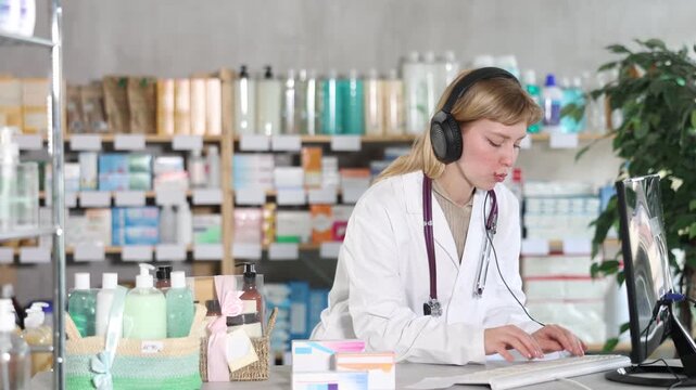 Attentive female pharmacist wearing headphones working with computer in chemist's shop. High quality 4k footage