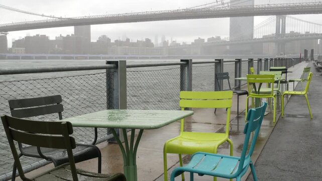 Brooklyn Bridge, Manhattan Bridge from Brooklyn Bridge park, New York City Dumbo United States. Waterfront promenade, riverside esplanade on rainy foggy day. Empty wet metallic chairs, tables by water