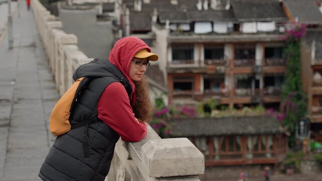 Asian woman leaning on bridge railing overlooking riverside village, wearing red hoodie, black puffer vest and yellow cap with orange backpack, contemplative posture, stone parapet, wooden balconies,