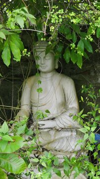 Two stone statues, one seated and one standing, are partially obscured by foliage in a garden in Ngu Hanh Son, Da Nang, Vietnam. The statues are weathered and covered in moss.