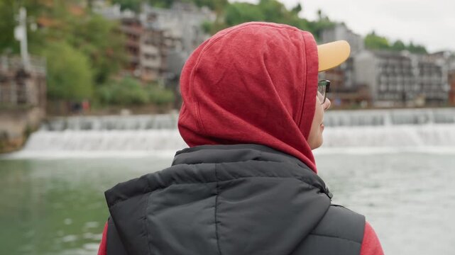 Young asian woman at riverside exploring, wearing red hoodie and yellow cap with orange backpack and glasses, glancing across waterfall toward wooden bridge and stilted houses, lively market energy,