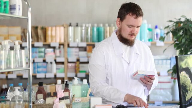 Young man pharmacist looking for box of pills on computer in pharmacy