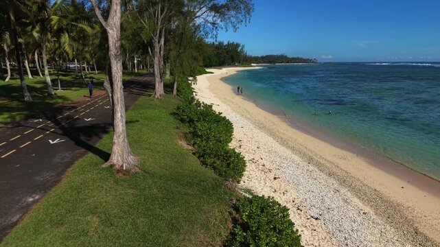 Aerial shot captures a tropical road lined with palm trees. Clear turquoise water meets a white sandy shore. Lush green forests border the coastline. No people are visible in the scene
