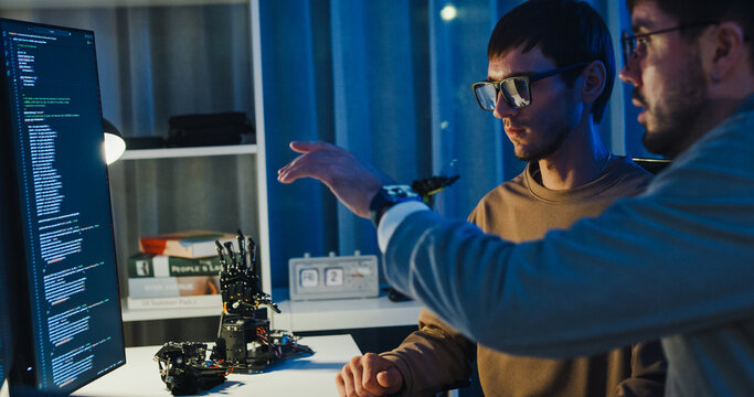 Team adult male engineer collaborate on robotic hand project using AI code solutions machine learning at tech workspace night. Artificial intelligence interface, collaboration prototype development.