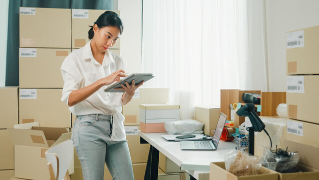 Youth attractive Asia female entrepreneur small business use digital tablet check parcel counting cardboard box in confirm check stock inventory product ready for shipment at small online retail shop.