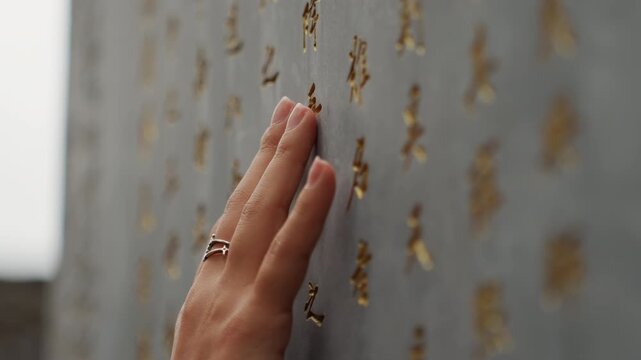 Closeup hand on engraved wall, detailed focus on fingers brushing gilded characters on weathered stone, soft overcast light, contemplative mood, tactile texture, slow cinematic movement
