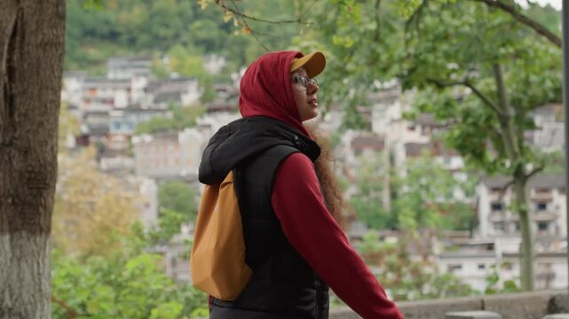 Woman with red hood and backpack walking past tree and lamp post, scanning terraced houses and street architecture, active explorer energy, casual cap and glasses, layered fall clothing and mustard