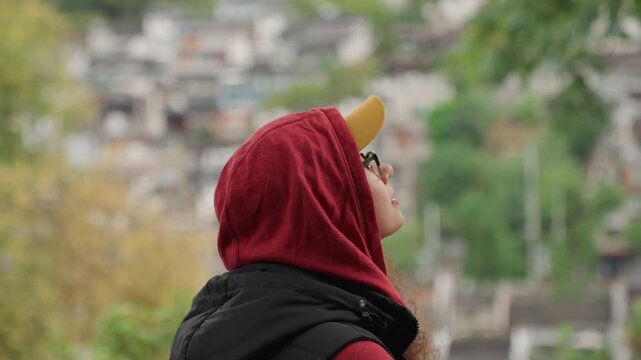 Young latina woman reaching toward tree, confident gesture toward branch, layered jacket and backpack, yellow cap and glasses, gaze over city valley, candid park portrait, autumn palette,