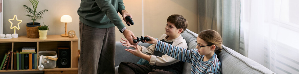 Fototapeta premium Caucasian boy and Caucasian girl sitting on sofa reaching for game controllers from adult man standing nearby, children preparing to play video game together in living room setting