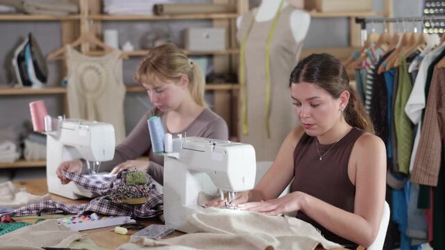 Two female tailors are sitting at a table in the workshop and sewing clothes on machines. Employees of the sewing industry make wear according to individual measurements. High quality 4k footage