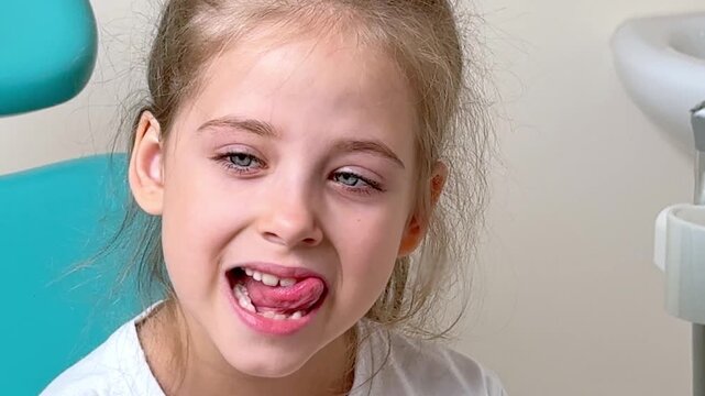 Little girl with light brown hair and blue eyes sits in a dental chair, sticking out her tongue during a dental examination, with dental tools and equipment visible in the background