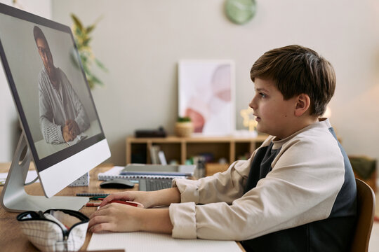 Caucasian boy sitting at desk participating in online lesson with young adult man on computer screen, focusing on monitor and interacting with digital learning materials in home environment