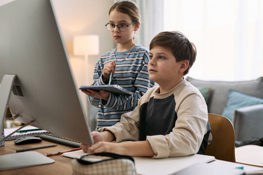 Caucasian boy sitting at desk using desktop computer while Caucasian girl standing beside holding digital tablet, both children focusing on screen in home environment