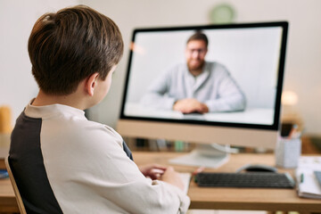 Fototapeta premium Caucasian boy sitting at desk watching middle aged Caucasian man on computer monitor during video call, boy facing screen with hands resting on table, home interior background visible