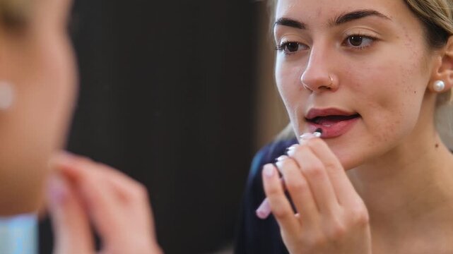 A woman applies a lip product using a small applicator while reflecting on her appearance in a mirror. This content is suitable for beauty, fashion, and wellness projects
