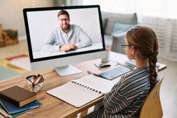 Fototapeta premium Caucasian girl sitting at desk participating in online lesson with Caucasian middle aged man on computer monitor, writing in notebook, wearing glasses, studying in home environment