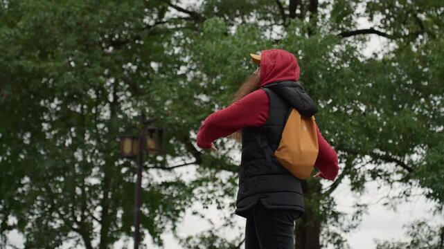 Young woman red hoodie performing outdoors, expressive gestures and animated arm movements, long hair, black puffer vest and yellow backpack, lamppost and leafy trees in overcast park, candid street