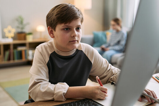 Caucasian boy child sitting at desk using desktop computer and writing in notebook, focused on screen, with another child blurred in background sitting on couch holding digital tablet
