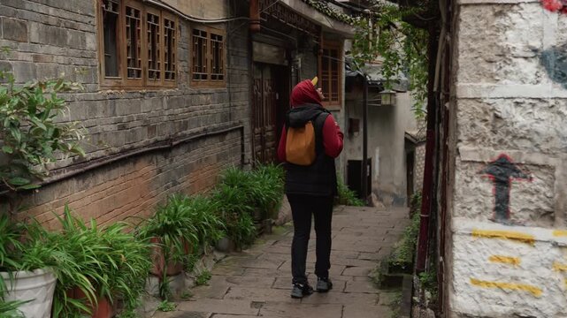 Red jacket orange backpack walking alley between stone buildings, narrow cobblestone path bordered by potted plants and wooden windows, overcast light, traveler exploring rustic village, reflective