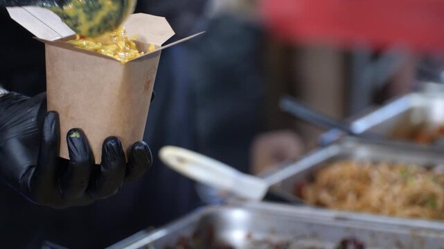 Close up of a vendor serving steaming hot dish from a professional warmer into a container. Authentic street food preparation and packaging for a client in a local canteen