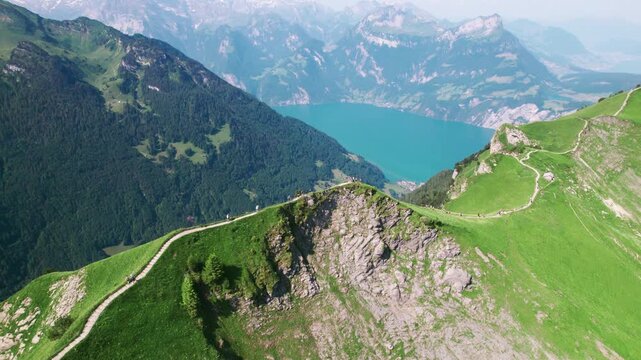 Alpine ridge landscape, winding mountain path, lush green hills, Swiss Alps panorama, turquoise lake below, summer hiking scenery, travel nature footage by 4k aerial drone.